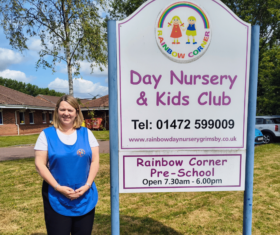 Jackie Woods standing next to the Rainbow Corner Day Nursery sign with the setting behind her. 